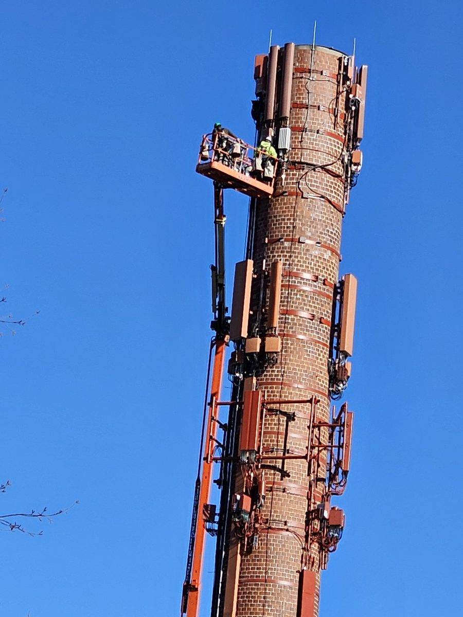 Two workers on a boom-lift platform working beside antennas mounted high on a brick-clad monopole tower