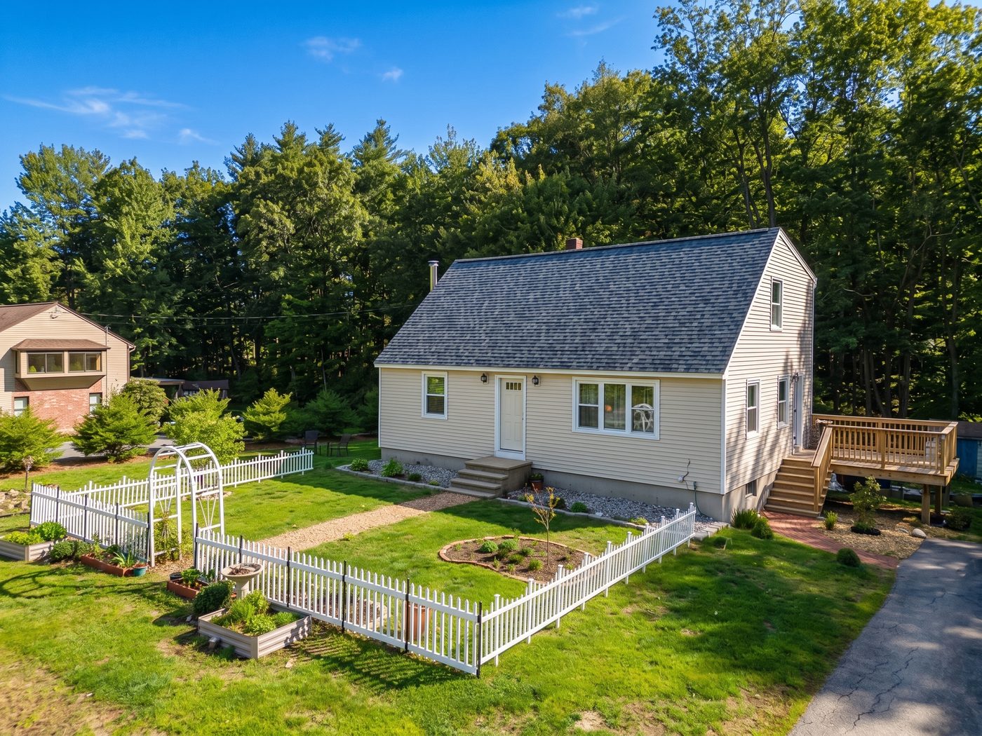 Aerial oblique of a cape-style house in summer sun, fenced garden beds and pine woods behind
