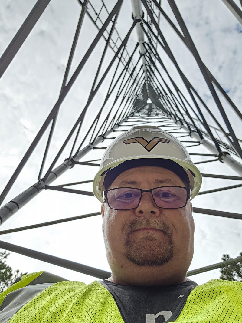 The pilot in a hard hat and high-visibility vest, standing at the base of a lattice tower that converges to a point far overhead