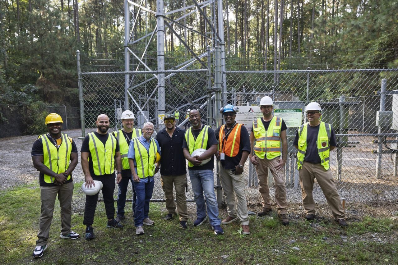 Nine people in high-visibility vests and hard hats standing together at the foot of a fenced telecom tower compound